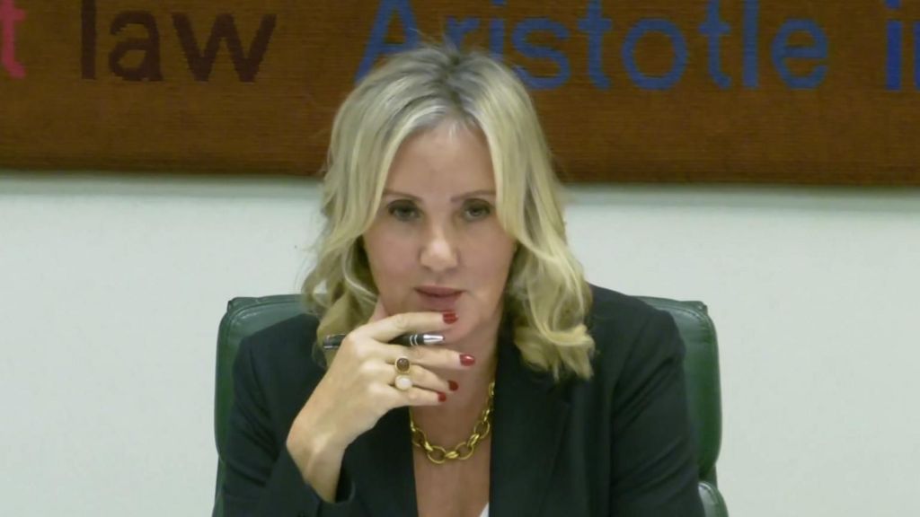 Caroline Dinenage, a woman with blonde waved hair, holds a pen and has her fingers on her chin - she is sitting in a dark green chair, and wears a dark jacket and a chunky gold necklace