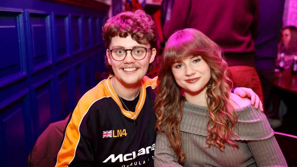 Kayleb and Alana smile at the camera in a pub. Kayleb has short curly hair and wears a racing jersey. Alana has long curled hair and wears a beige jumper.
