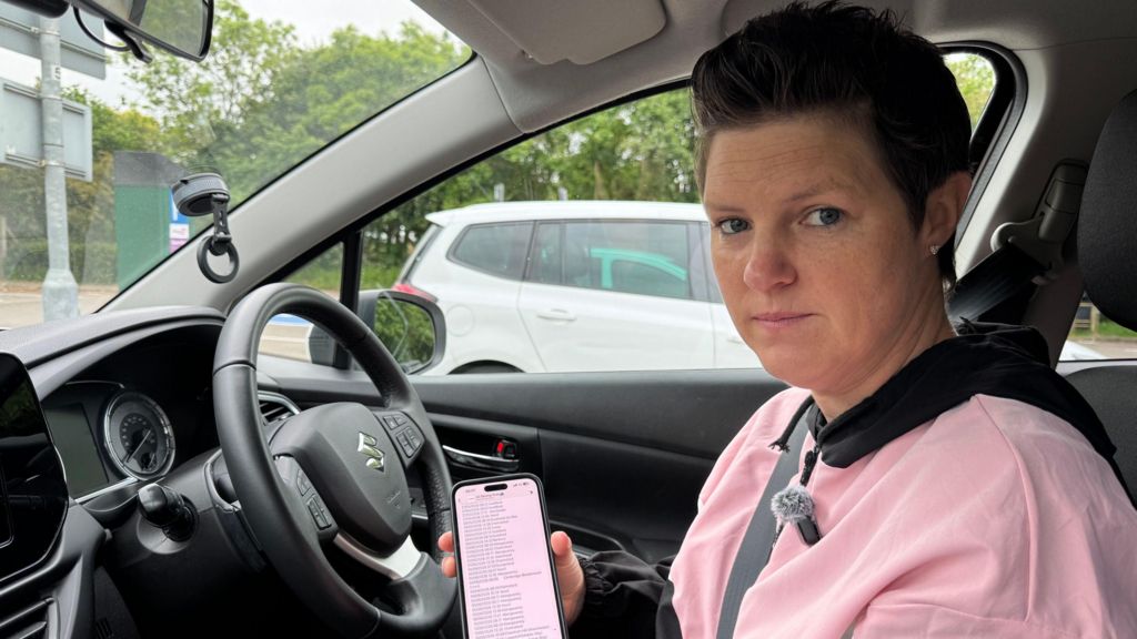 Sophie, a white British woman sits at the steering wheel of her car that she uses for driving lessons. She has very short brown hair and wears a pink tracksuit top and is holding her phone to the camera showing driving tests for sale.