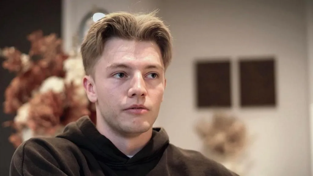 A young man with middle parted, cropped light blonde hair looks across the frame. He is wearing a black hoodie and is sat in front of a vase of flowers in an indoor area.