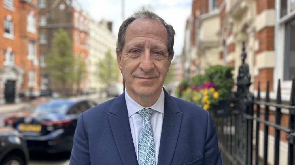 Jeremy wears a grey-blue suit jacket, white shirt and blue patterned tie while standing on a street in London.