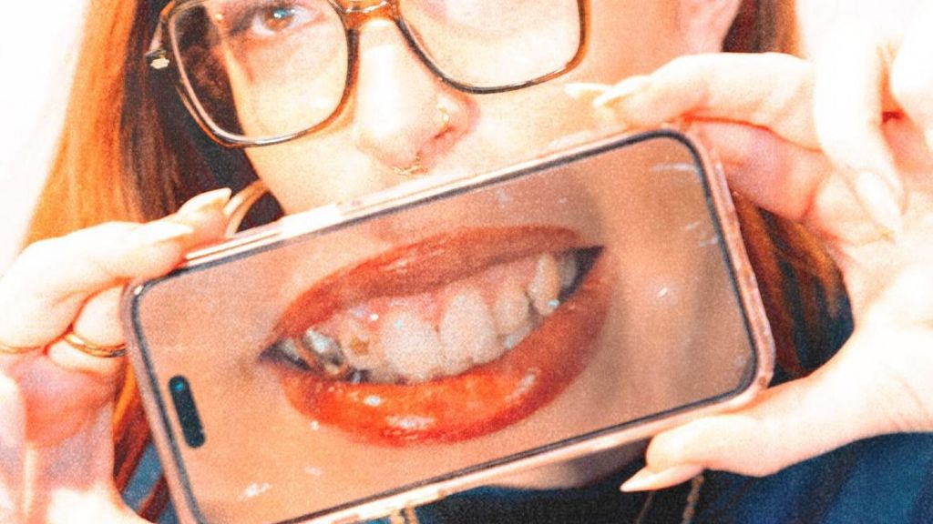 A woman with ginger hair and glasses holds a phone up against the lower half of her face. The phone shows a picture of her smiling with her teeth as she shows off her tooth gems.