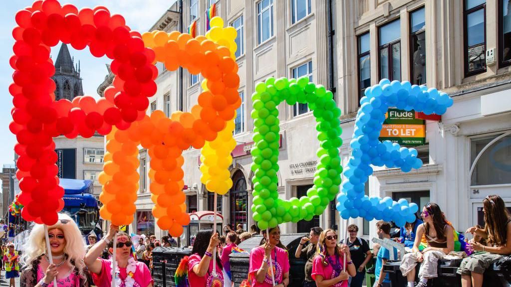 A drag queen and four ladies in pink tops walk down the street holding a different letter of the word pride and each in a different colour