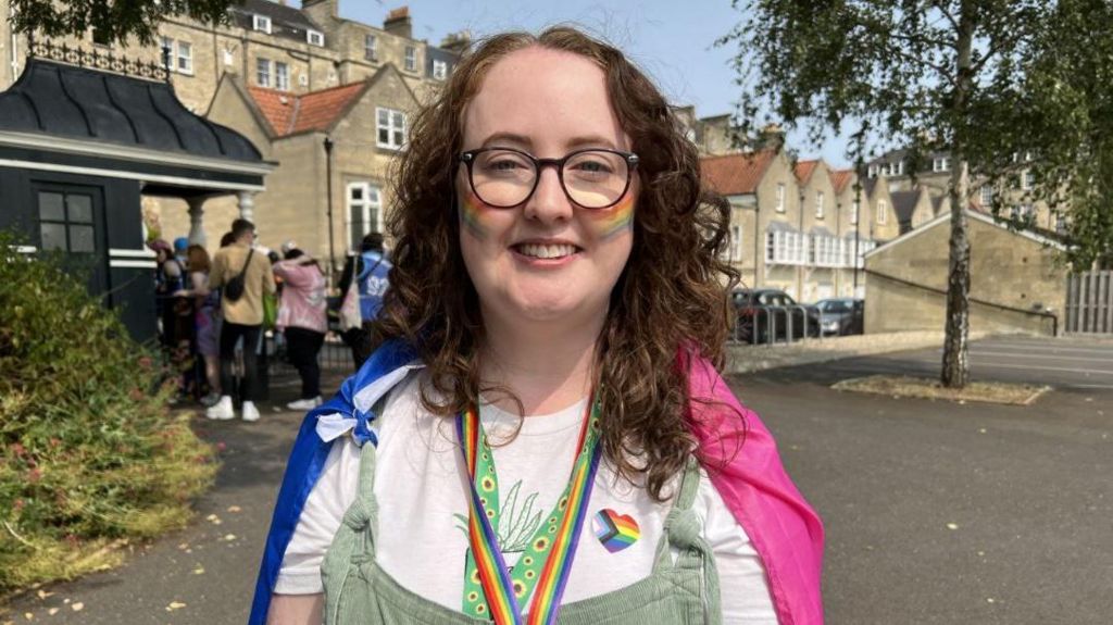 Claire Warner who has brown curly hair and glasses. She is wearing green dungarees and a Pride flag is draped on her shoulders. She also has the Pride flag painted on her cheeks.