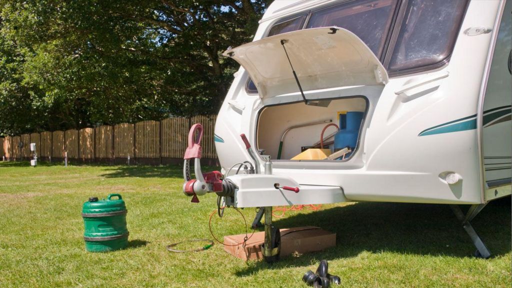 A caravan close up with it's boot open and trailer visible. It's parked on grass on a sunny day with a gas canister nearby