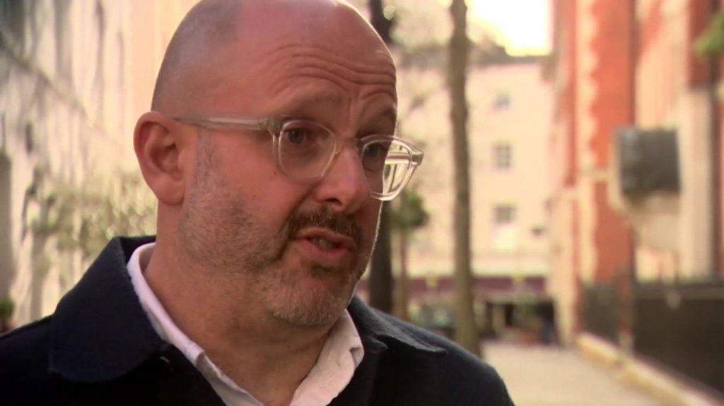 A middle aged man with glasses and a beard being interviewed for TV, he is wearing a dark coat and open-necked pink shirt. 