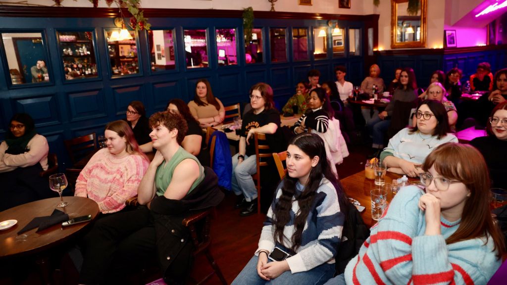 A crowd shot of the pub, which shows several tables with people sat around them, watching a TV screen out of shot. The crowd is mostly women, and most are young.