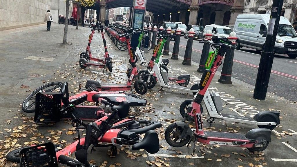 A group of e-scooters and e-bikes on the pavement. Some of them have fallen on the floor, others are not in the marked parking bay. 