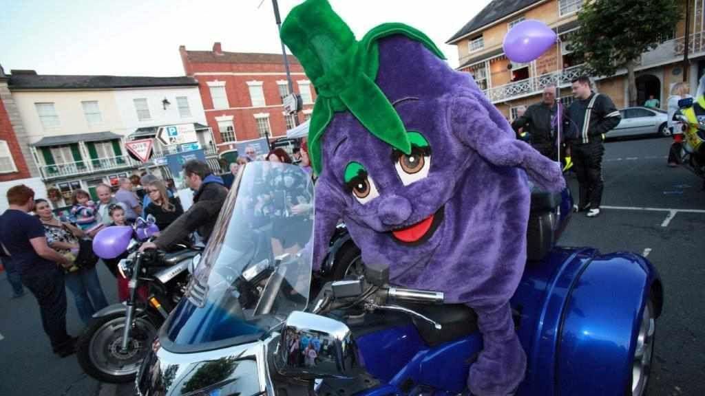 A purple cartoon character costume representing a plum with arms and legs is sat on a blue motorbike