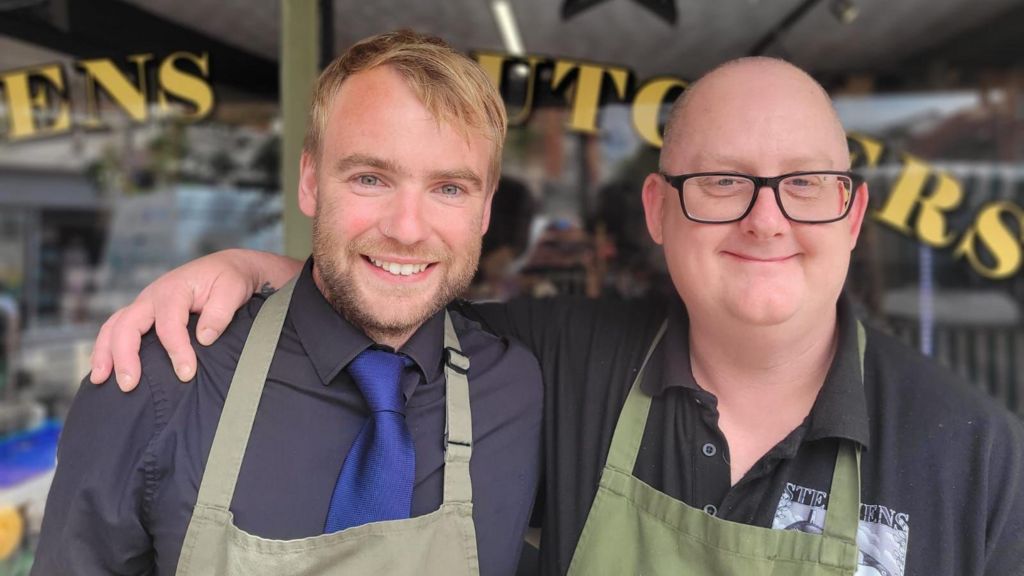 Two men are standing in front of a window to a butchers with their arms around each other. Both are wearing black shirts and green aprons.