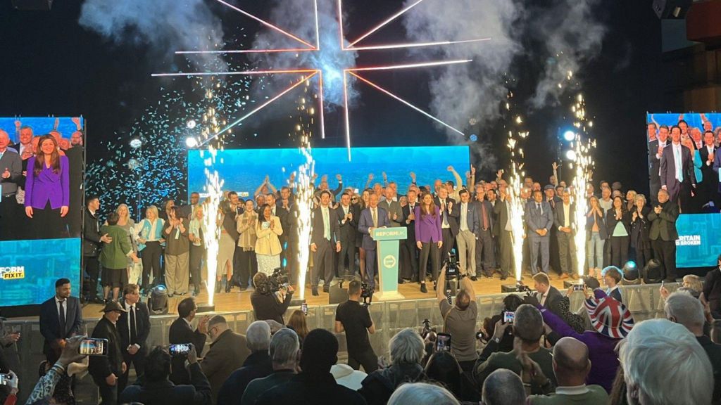 A big crowd of people on a stage, with Nigel Farage in the middle, all wearing suits and formal wear. There are four big sparklers at the front of the stage and an audience.