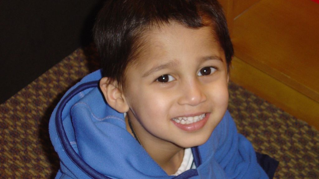 A young Amrit at 4-years-old. He's sat looking up and smiling at the camera wearing a blue jumper.