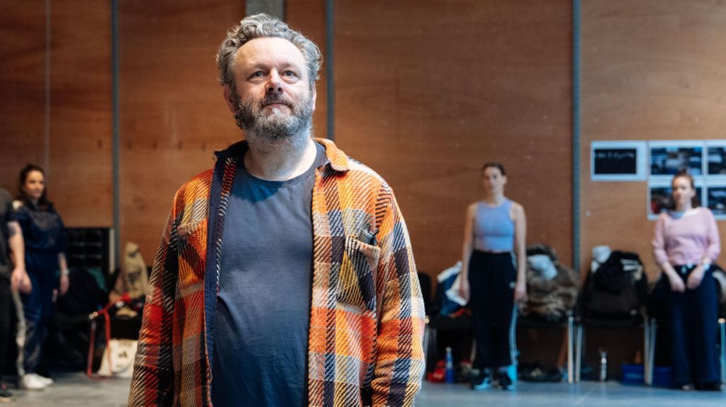 Michael Sheen in a checked shirt and T-shirt in the foreground, with other actors at the back of the rehearsal room