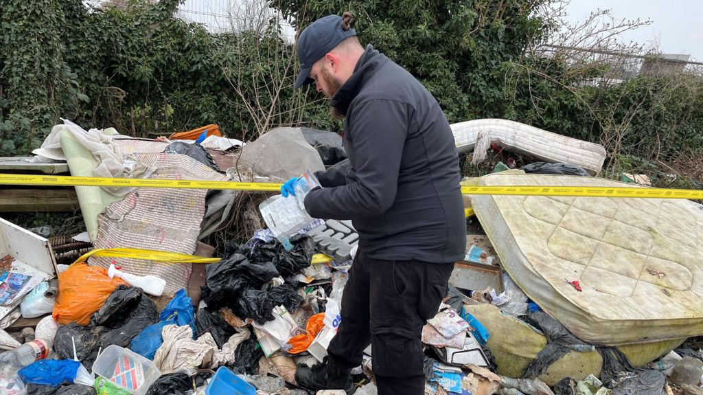 A man wearing a black cap, fleece-type top and trousers with blue plastic gloves is searching through rubbish including bin bags and discarded dirty mattresses