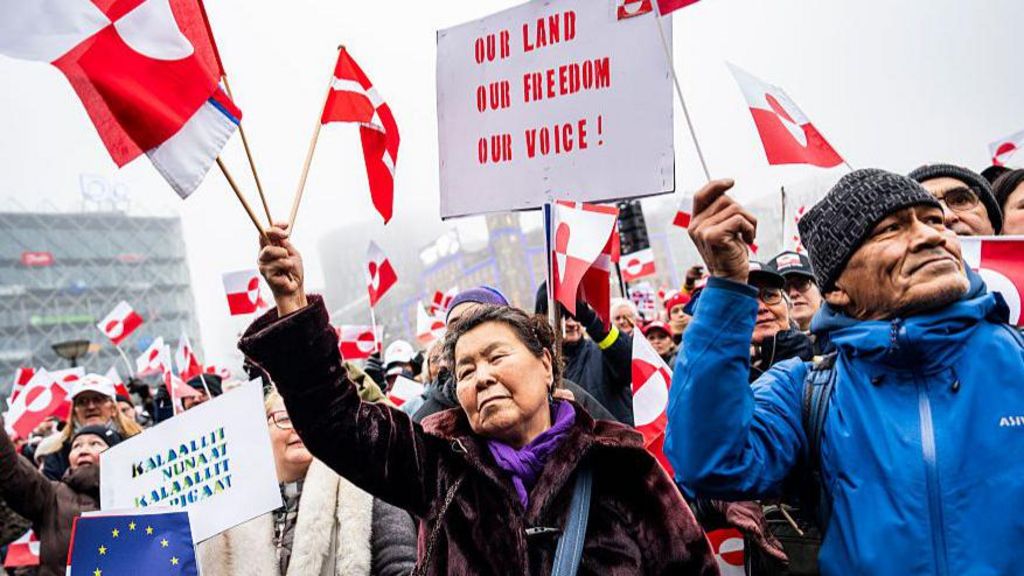 A crowd of protesters holding red and white Greenlandic flags and signs rally at City Square in Copenhagen, Denmark. They are dressed in winter coats, with one man wearing a beanie. A sign at the top of the image reads: "Our land, our freedom, our voice!"