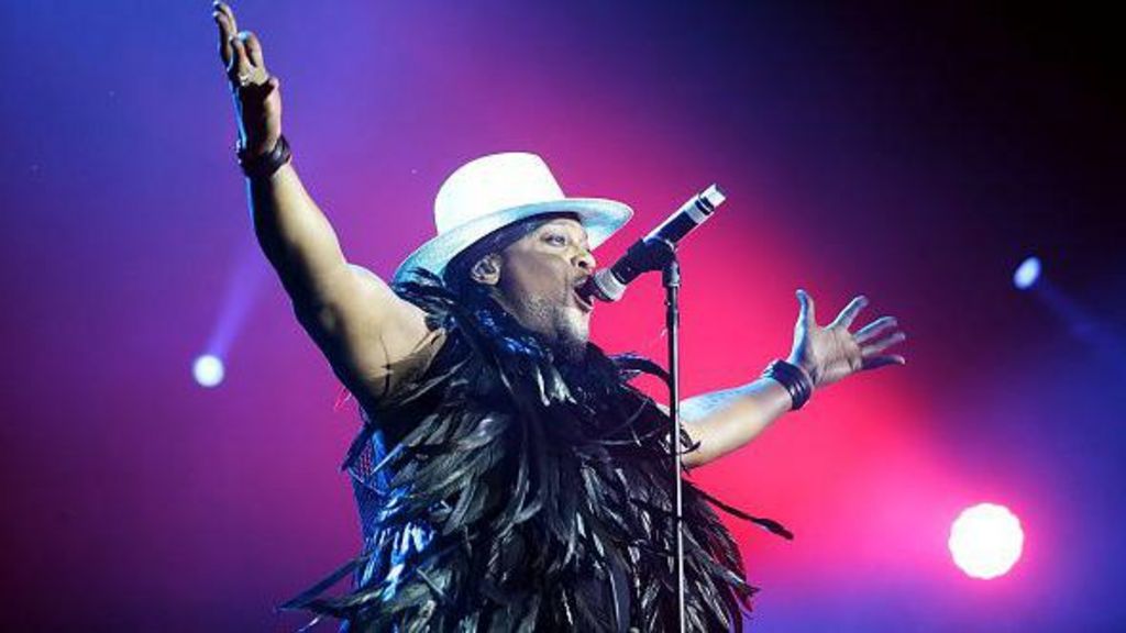 D'Angelo, wearing a white hat and a boa with black feathers around his neck, performs on a stage with bright pink lighting. He is raising his arms in the air. 