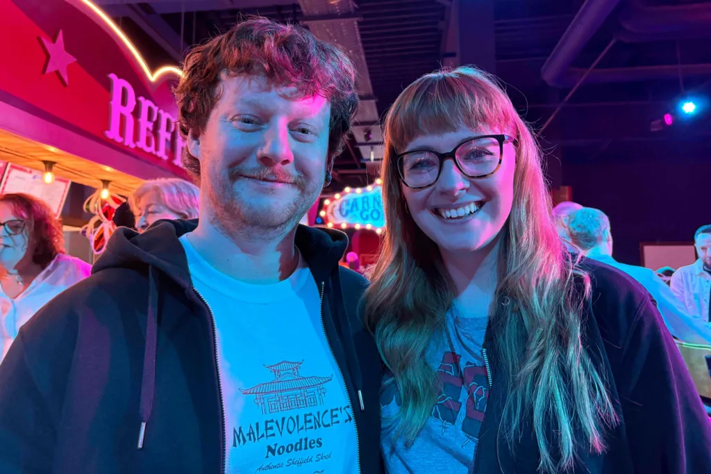 A smiling Jake and Lily Saunders. He is wearing a dark hoodie and white tee-shirt. She has long red hair and is wearing glasses, a dark hoodie and grey tee-shirt