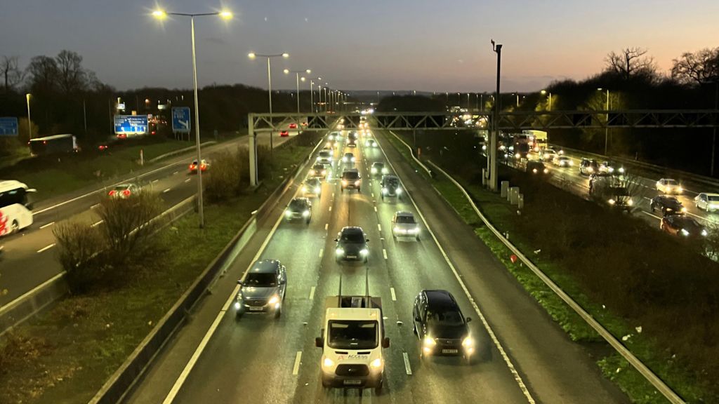 Cars with their headlights on drive towards a bridge on the triple-laned M1 motorway as the sun sets.