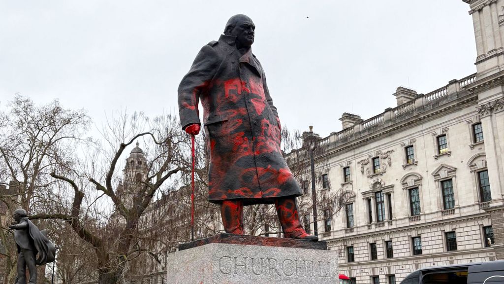 Winston Churchill statue seen from the left front side, looking up at the statue,  covered in red paint. Buildings in the background 