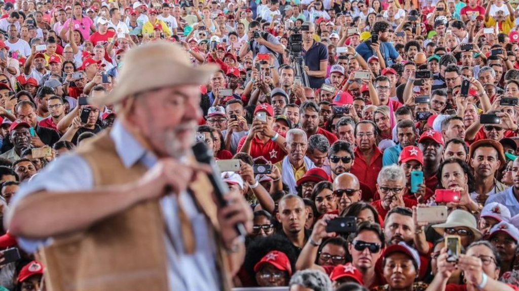 Lula discursa em cima de palco e é observado pela plateia, onde há diversas pessoas com bonés ou camisetas do PT