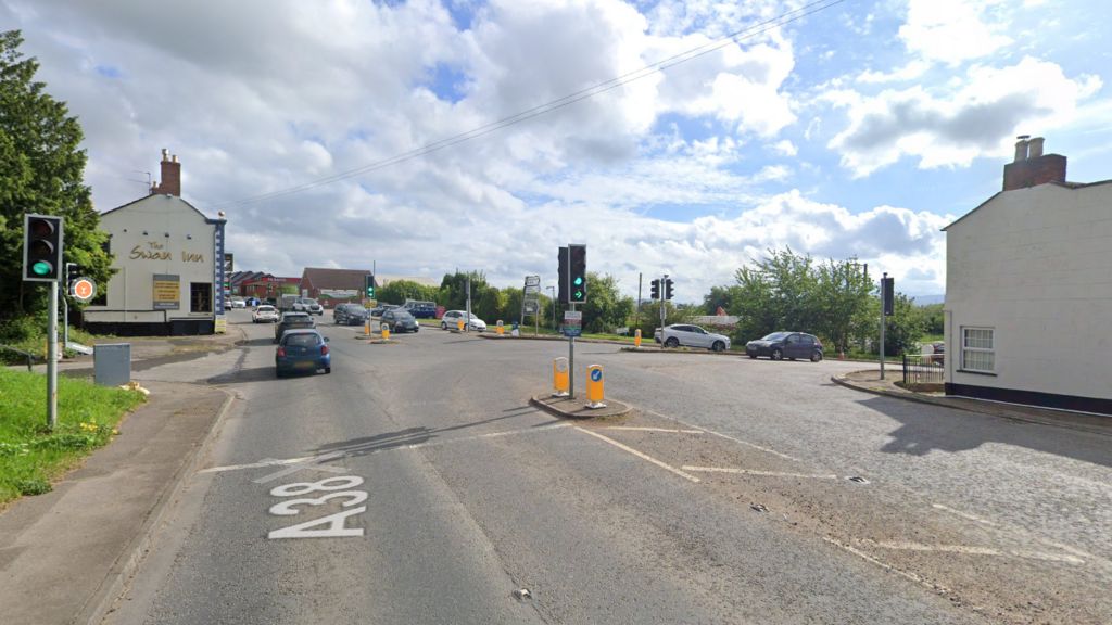 A section of the A38 with traffic lights and cars passing through. On the left, the side of The Swan Inn pub can be seen.