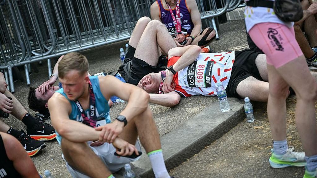 Several marathon finishers sit or lie flat on the ground in a designated rest area, showing visible signs of fatigue after completing the race.