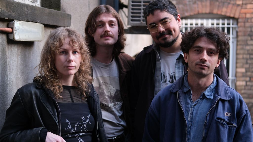 Four members of Brown Horse standing together and looking at the camera outdoors in front of brick and concrete walls.