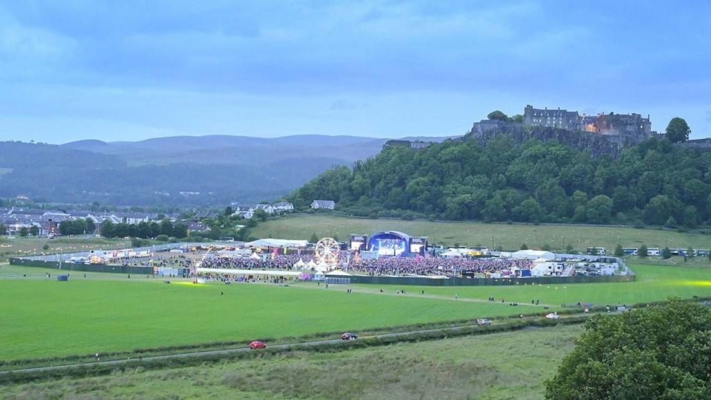 A large park with a stage in the middle and crowds. Stirling Castle is visible in the background