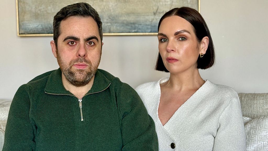 Couple pictured sitting on a sofa. John, on the left, has short, dark hair and a beard. He is wearing a ribbed green jumper with a zipped neck. Katie has straight, dark, bobbed hair. She is wearing a v-necked white jacket fastened with a gold button. The sofa is cream and there is a piece of abstract art on the wall.