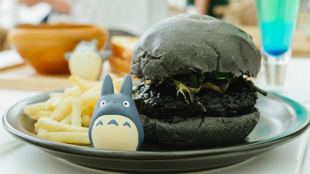 A black burger, on a plate with a small Totoro model, and fries