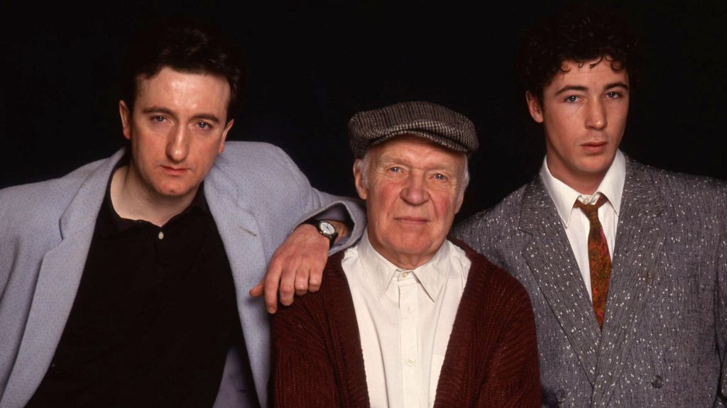 Three men pose together against a dark background. An older man in a flat cap and red cardigan stands in the center with his arms folded, while two younger men stand on either side—one leaning in with his arm on the older man’s shoulder, and the other in a gray suit. All three look directly at the camera.