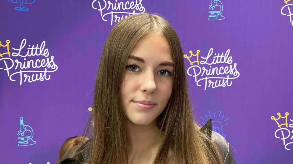 Une jeune fille aux longs cheveux bruns pose devant un fond violet orné du logo du Little Princess Trust.
