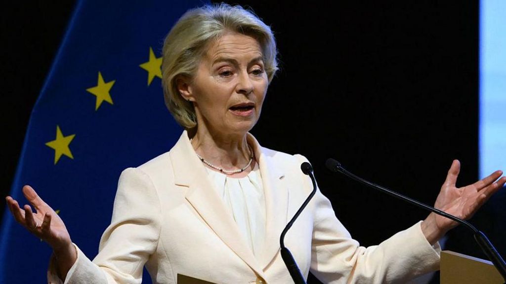 European Commission President Ursula von der Leyen wearing cream suit stands in front of the European Union flag
