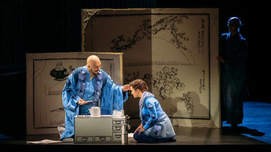 A man and a woman wearing blue robes kneel on the stage. There is a small kitchen unit on the stage and two boards in the background with black ink drawings, inspired by Japan.