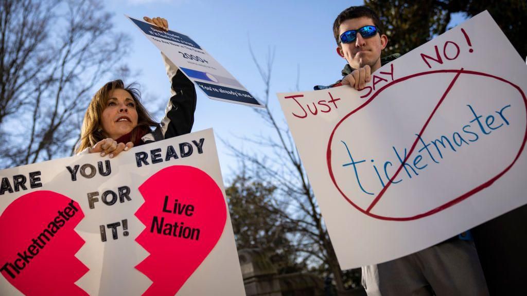 Taylor Swift fans outside the US Capitol, holding signs showing a heart split in two with "Ticketmaster" and "Live Nation" written on each half, along with the slogan "Are you ready for it?", a reference to a Taylor Swift song; and another sign with "Ticketmaster" in a red circle with a line through it, and the slogan "Just say no!" 