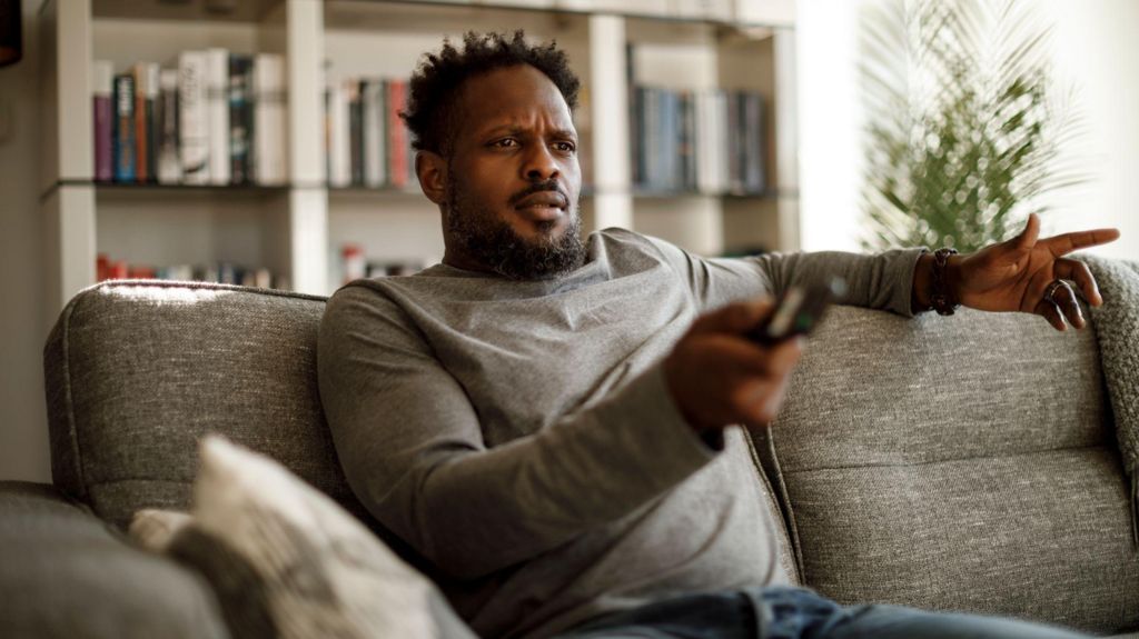 Man sitting on a sofa holding a TV remote with a confused expression