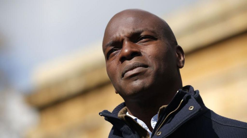 A close-up of Lord Bailey wearing a dark coat, standing outdoors in bright sunlight with a blurred stone building in the background.