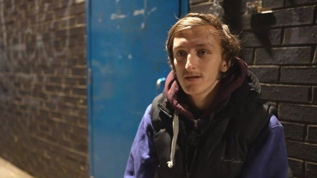 Samuel Atkinson sitting outside a homeless shelter. He is wearing a purple jumper and black gilet.