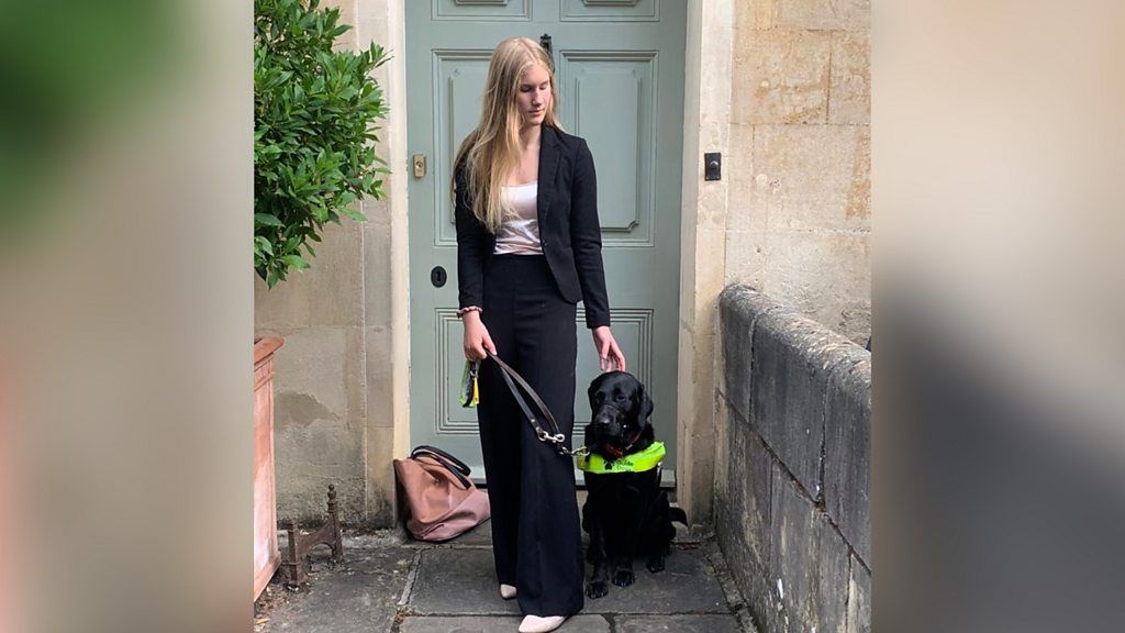 A teenage girl stands in front of a front door with a guide dog on a lead who is sitting down