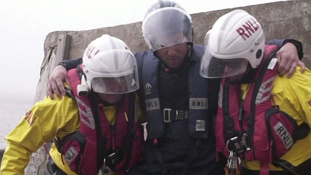 In a reconstruction, two RNLI officers help an injured man