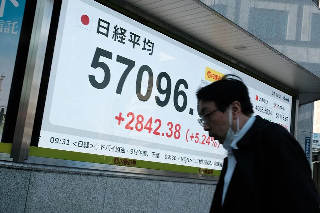 A man walks in front of a giant screen showing the Nikkei Index at 57096