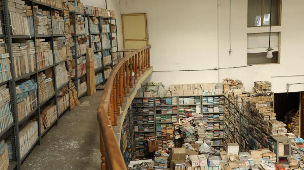 The photo shows part of the upper floor of a building with books stacked neatly on shelves. On the lower floor, there are hundreds of books on the shelves and piled up on the floor.