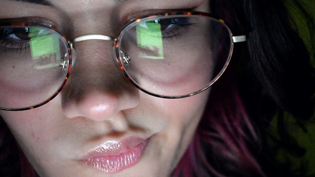 A picture of a young woman wearing glasses, in the reflection you can see a mobile phone
