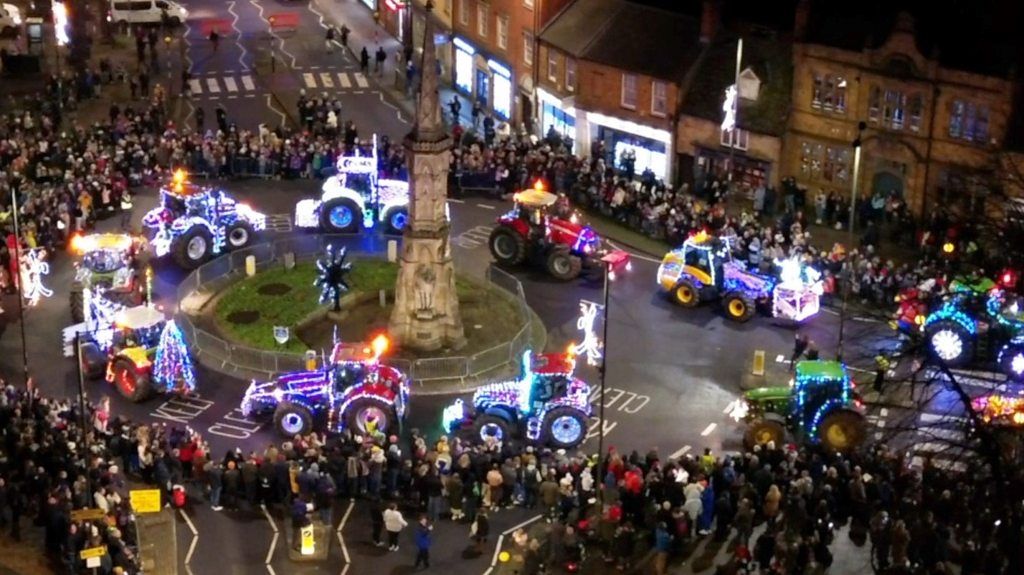 Tractors illuminated with Christmas lights drive through a town