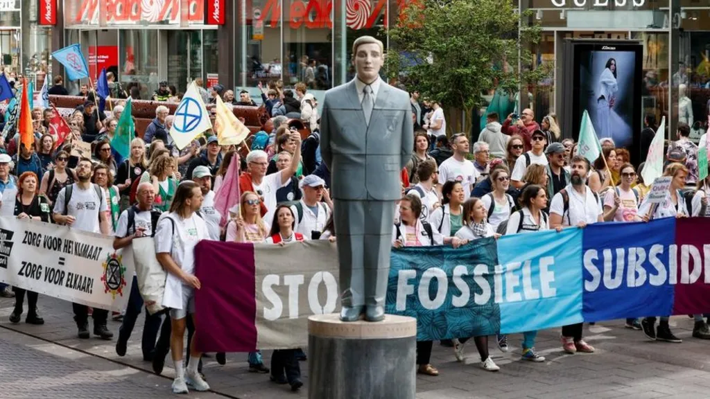 The image shows a climate protest with a large crowd of demonstrators gathered around a statue of a man in a business suit on a pedestal. Protesters are holding banners with text including 