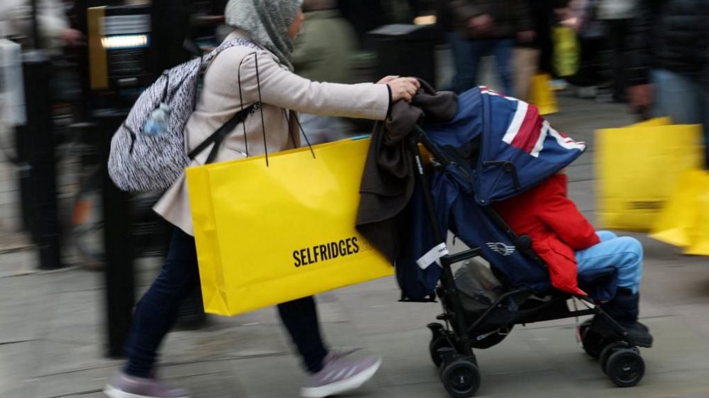 A woman wearing a hijab pushes a pram while carrying a large yellow Selfridges bag down the street.