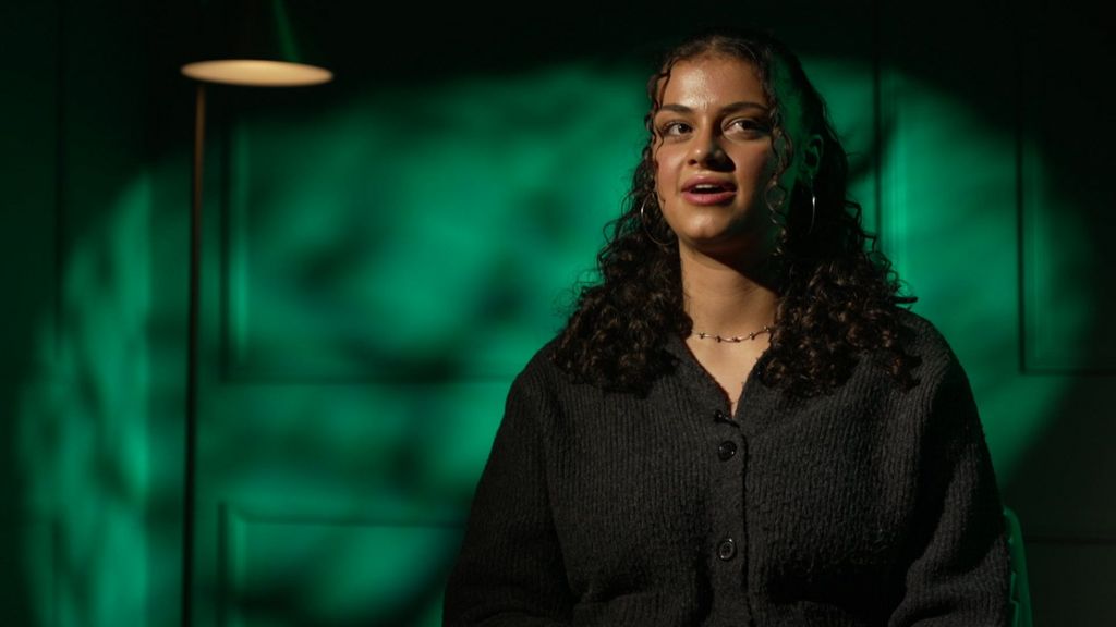 Ruby Greensides sits facing the camera in a studio interview, wearing a dark cardigan, with green light patterns behind her.