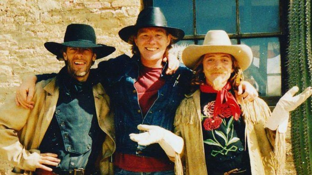 Three men wearing cowboy hats and smiling at the camera. The one on the right is Rik Mayall. The two leftmost men are wearing black hats, while Mayall is wearing a white one. The men on the far left and far right are wearing other cowboy clothing.