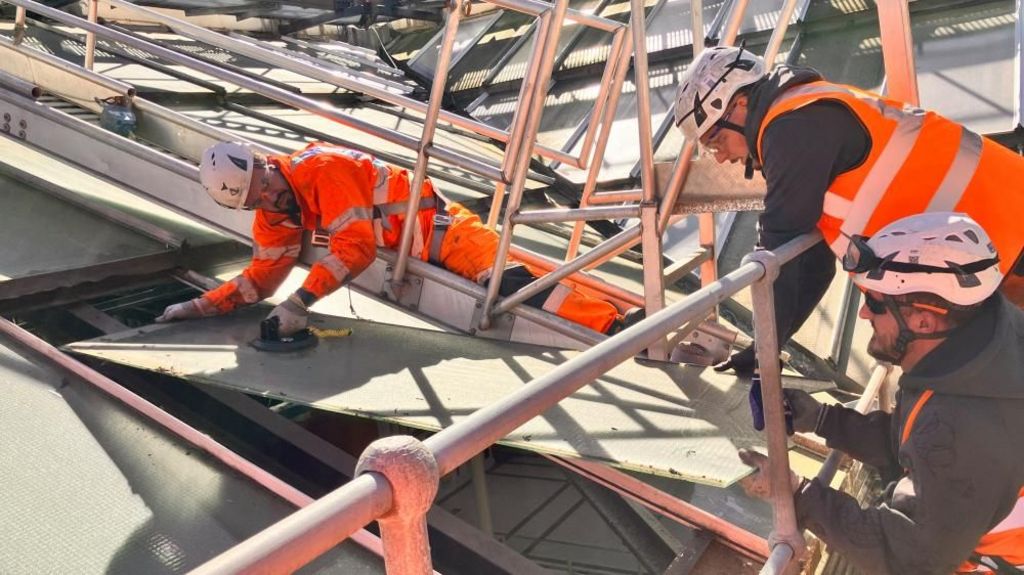Three men in high visibility orange jackets work and helmets work on the roof installing a new panel.