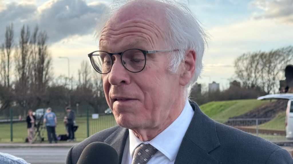 Nic Dakin in a dark suit and patterned tie stands outdoors by a roadside, speaking into a handheld microphone. A van, grassy embankment, and distant figures are visible in the background under a cloudy sky, suggesting a media interview taking place.